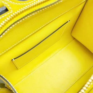 May include: The interior of a bright yellow handbag featuring a smooth leather lining, white contrast stitching along the seams, and a single flat open pocket attached to the back wall. The bag has a structured shape with visible edge piping.