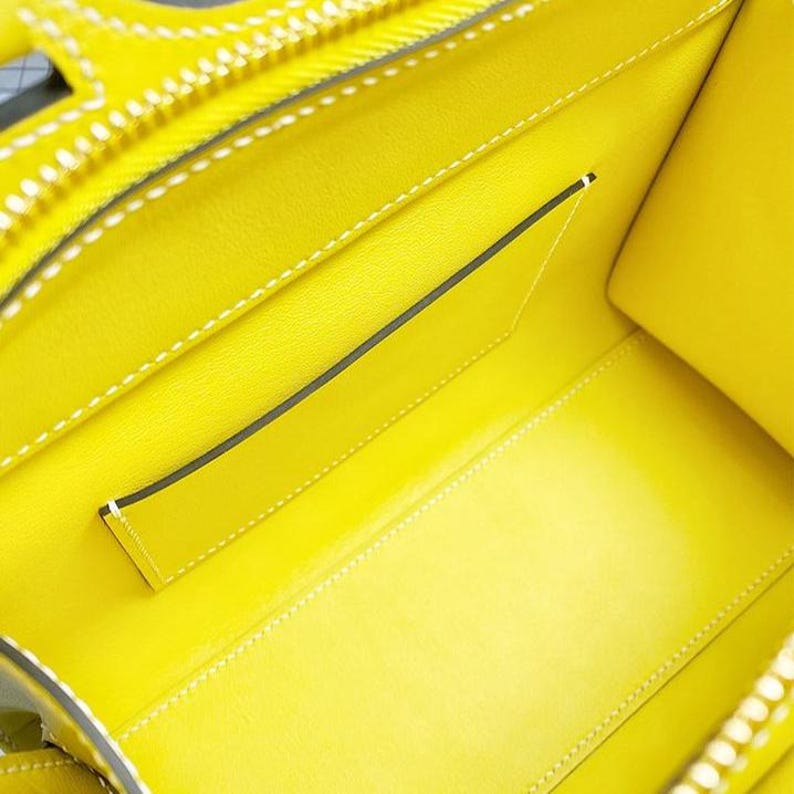 May include: The interior of a bright yellow handbag featuring a smooth leather lining, white contrast stitching along the seams, and a single flat open pocket attached to the back wall. The bag has a structured shape with visible edge piping.