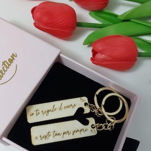 May include: A set of two wooden keychains with a gold chain connecting them. The keychains are engraved with the text "io ti regalo il cuore..." and "e resto tua per sempre". The keychains are in a pink box with red tulips.