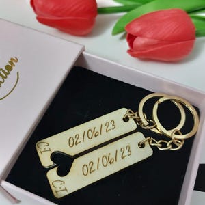 Puede incluir: Un llavero de madera con dos etiquetas, cada una grabada con la fecha "02/06/23". Las etiquetas están conectadas por una cadena dorada y un anillo dorado. El llavero está en una caja rosa con interior negro.