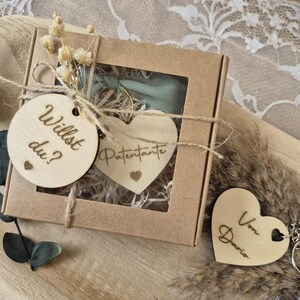 Könnte beinhalten: Eine Geschenkbox mit einem herzförmigen Holzschlüsselanhänger und zwei Holzanhängern. Auf den Anhängern stehen die Aufschriften "Willst du?" und "Patentante". Die Box ist mit Bindfaden gebunden und mit Trockenblumen verziert.