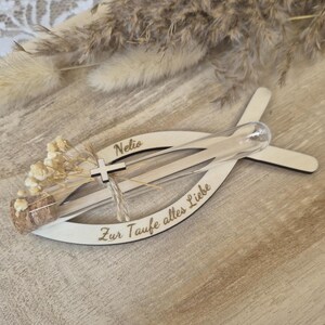 May include: A light-colored wooden fish-shaped ornament featuring the name Nelio and the German text Zur Taufe alles Liebe. A clear glass test tube with a cork stopper is attached, containing small dried flowers and a tiny wooden cross charm.