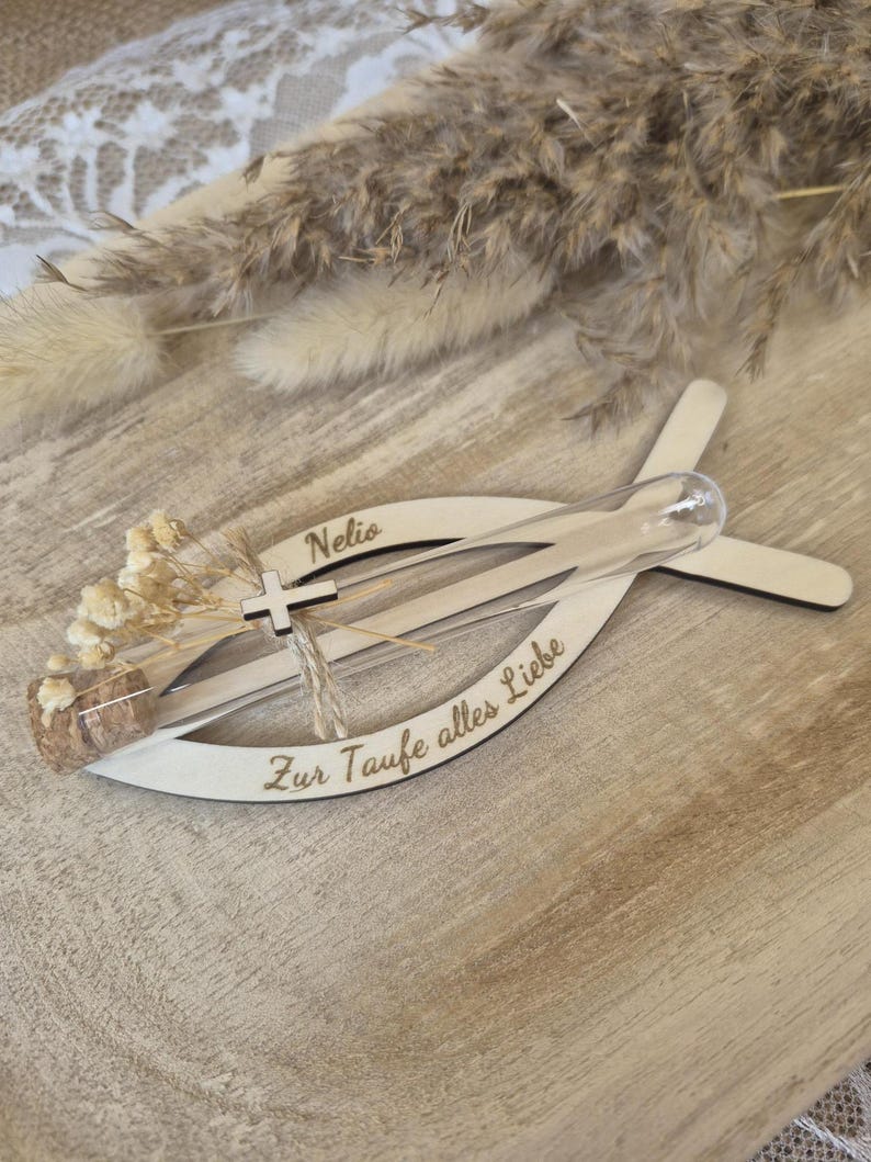 May include: A light-colored wooden fish-shaped ornament featuring the name Nelio and the German text Zur Taufe alles Liebe. A clear glass test tube with a cork stopper is attached, containing small dried flowers and a tiny wooden cross charm.
