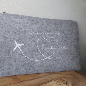Könnte beinhalten: Graue Filz-Reisemappe mit weißen Nähten. Die Mappe hat ein weißes Flugzeug mit einem gepunkteten Linienweg, der zu einem Herzen mit dem Text "Reisedokumente von Familie Votta" führt.