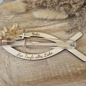 May include: A fish-shaped wooden cutout featuring the engraved name Nelio and the German text Zur Taufe alles Liebe. A clear glass test tube with a cork stopper is attached, decorated with a small wooden cross and dried cream-colored flowers.
