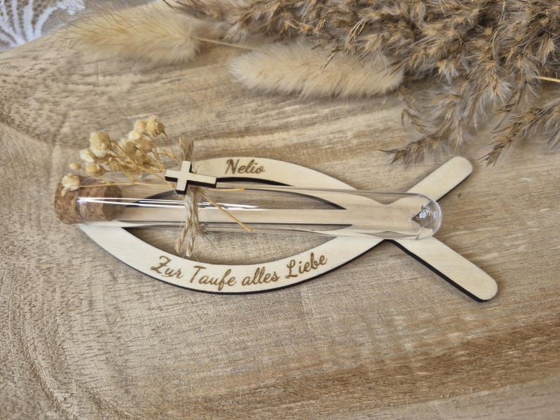 May include: A fish-shaped wooden cutout featuring the engraved name Nelio and the German text Zur Taufe alles Liebe. A clear glass test tube with a cork stopper is attached, decorated with a small wooden cross and dried cream-colored flowers.