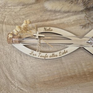 May include: A fish-shaped wooden plaque featuring the name Nelio and the German text Zur Taufe alles Liebe. A clear glass test tube with a cork stopper is attached, containing dried white flowers and a small silver-colored cross charm.