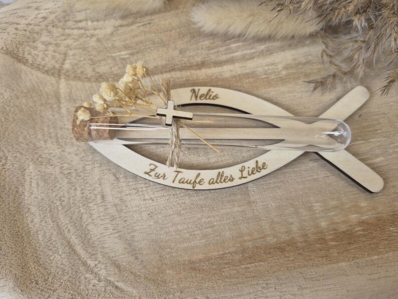 May include: A fish-shaped wooden plaque featuring the name Nelio and the German text Zur Taufe alles Liebe. A clear glass test tube with a cork stopper is attached, containing dried white flowers and a small silver-colored cross charm.