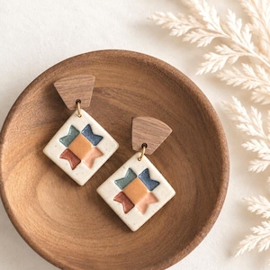 May include: A pair of dangle earrings featuring a dark wood fan-shaped top piece connected to a diamond-shaped white ceramic pendant. The pendant displays a central orange square surrounded by four star-like points in blue, pink, and rust colors.