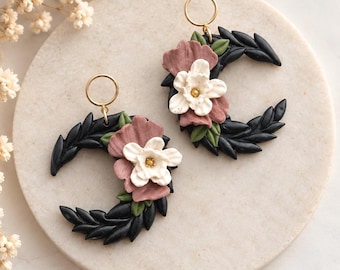 Op de afbeelding: Een paar oorbellen in de vorm van een halve maan met een zwart frame met bladstructuur, versierd met gebeeldhouwde oudroze en witte bloemen, geaccentueerd met kleine groene blaadjes en afgewerkt met een goudkleurige ronde metalen ring.