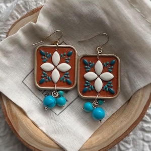 May include: A pair of square earrings with a floral design. The earrings feature a rust-colored background with white and teal accents. Turquoise beads dangle from the bottom. The earrings are displayed on a wooden surface with a white cloth.