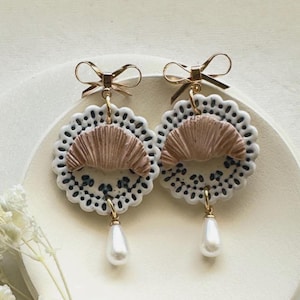 May include: A pair of handcrafted earrings featuring a circular design with a scalloped edge. The earrings have a tan, fan-shaped center, black and white dotted details, and a faux pearl drop. Gold-colored bows top the earrings.