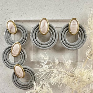 May include: A pair of gray and white statement earrings with a gold accent. The earrings feature three concentric circles with a pink and white oval stone in the center.