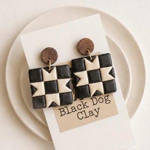 May include: A pair of square earrings featuring a black and cream geometric star pattern. Each earring is attached to a small circular wooden stud. The earrings are displayed on a white card with the text Black Dog Clay printed in black ink.