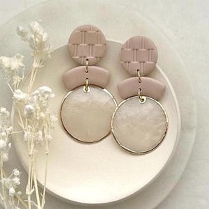 May include: A pair of beige and gold earrings. The earrings feature a textured circle at the top, a small rectangular shape in the middle, and a large, round, gold-rimmed circle at the bottom. The earrings are displayed on a white, layered plate with dried white flowers.