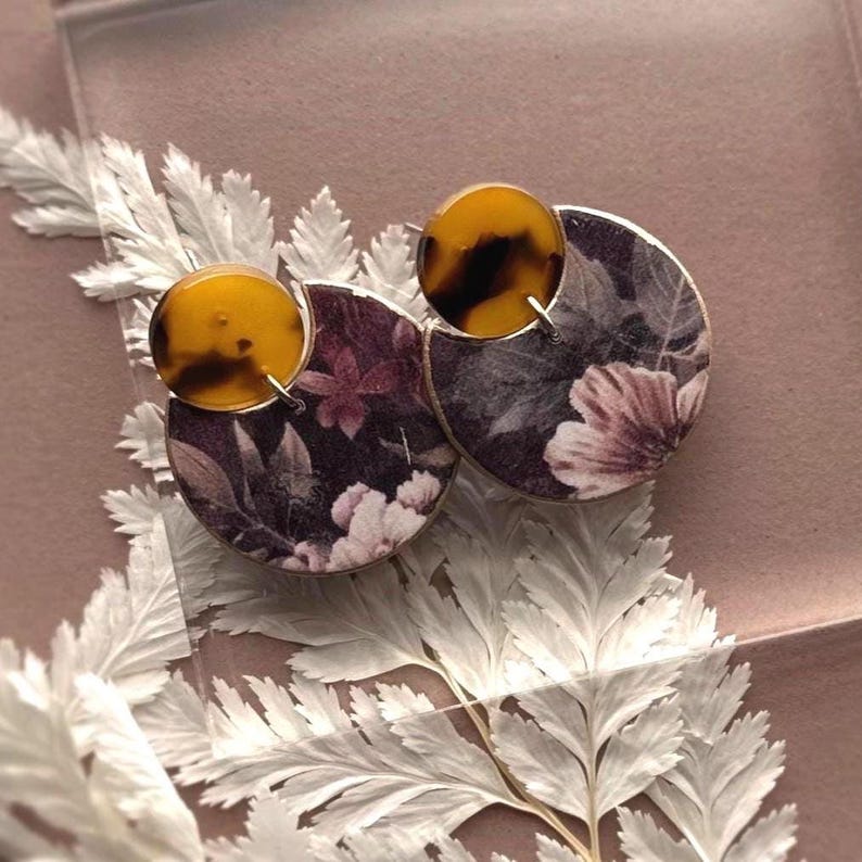 May include: A pair of floral earrings. Each earring features a half-circle design with a dark background and a floral pattern in shades of pink and white. A round, tortoiseshell-patterned accent sits atop each half-circle, creating a unique and stylish accessory.