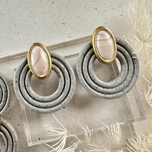 May include: A pair of gray ceramic earrings with a pink and white oval-shaped stone in the center. The earrings have three concentric circles.