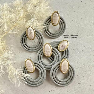 May include: Four pairs of gray ceramic earrings with a gold ring and a light pink oval stone in the center. Each earring is 1.2 inches long and 1.2 inches wide.