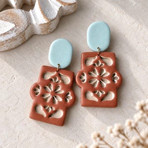 May include: A pair of dangle earrings featuring a light blue oval top piece connected to a rectangular terracotta-colored base. The terracotta base has a cutout floral pattern that reveals a lighter beige interior layer, creating a contrast.