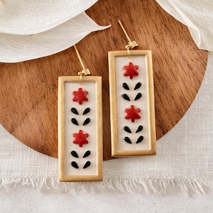 May include: A pair of rectangular drop earrings featuring a gold-toned frame and a white center inlay. The design includes two red six-pointed star shapes and four black teardrop-shaped accents arranged in a vertical pattern on each earring.