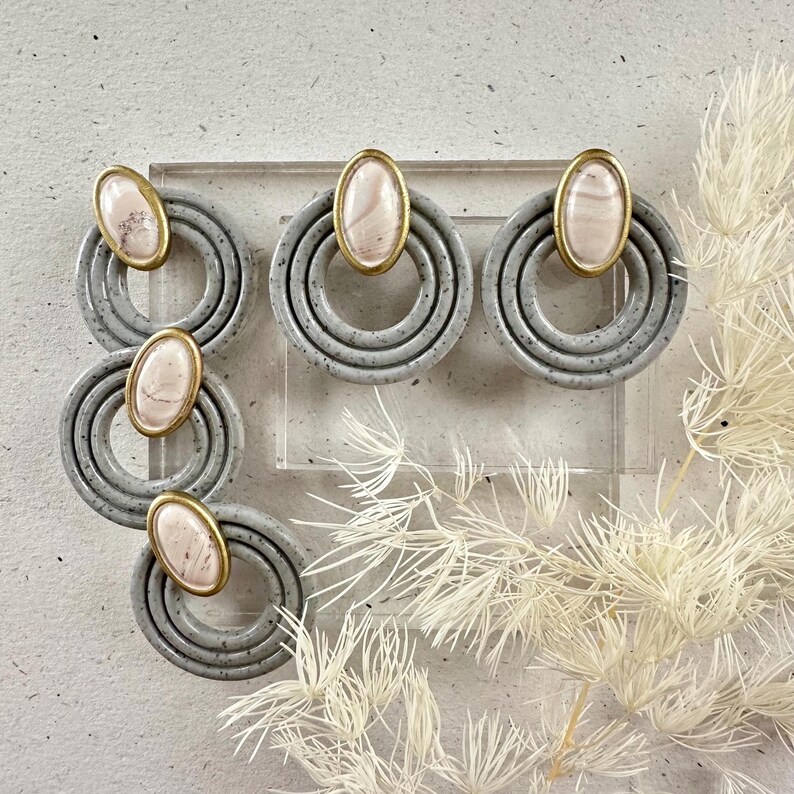 May include: A pair of gray and white statement earrings with a gold accent. The earrings feature three concentric circles with a pink and white oval stone in the center.