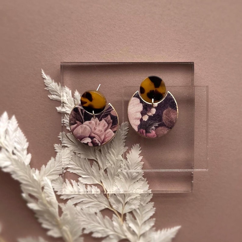 May include: A pair of earrings with a floral design. Each earring has a tortoiseshell-patterned top and a half-circle bottom with a floral print in shades of pink, purple, and white. The earrings are displayed on a clear acrylic stand with white decorative foliage.