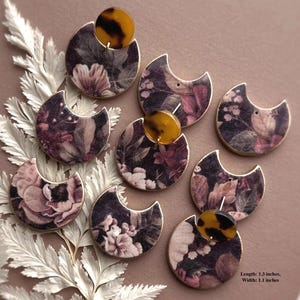 May include: A collection of floral print earrings. Each earring features a crescent shape with a dark background and a floral pattern in shades of pink, purple, and white. A round, tortoiseshell-patterned accent is attached to each earring. The earrings are 1.3 inches long and 1.1 inches wide.