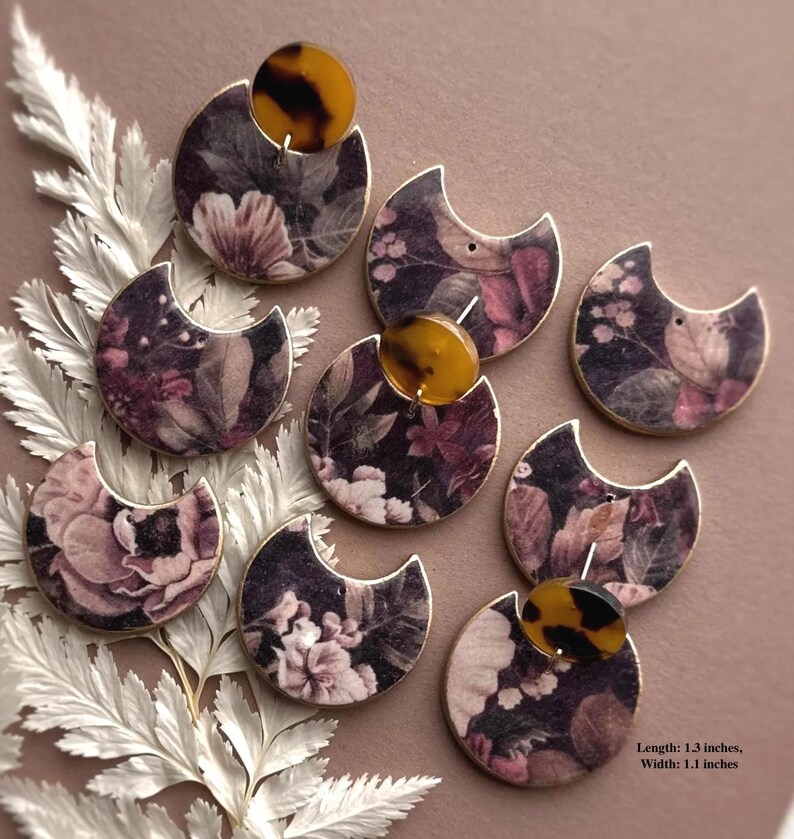 May include: A collection of floral print earrings. Each earring features a crescent shape with a dark background and a floral pattern in shades of pink, purple, and white. A round, tortoiseshell-patterned accent is attached to each earring. The earrings are 1.3 inches long and 1.1 inches wide.