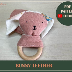 Op de afbeelding: Een roze konijnen-bijtring met een houten ring. Het konijn heeft witte oren en een bloemenpatroon op de rug. De bijtring is een geweldige manier voor baby's om hun tandvlees te kalmeren.