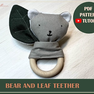 Peut inclure: Un hochet en tissu gris et vert en forme d'ours avec un anneau en bois. L'ours a une oreille en forme de feuille et un visage brodé simple. Le hochet est un excellent moyen pour les bébés de soulager leurs gencives.