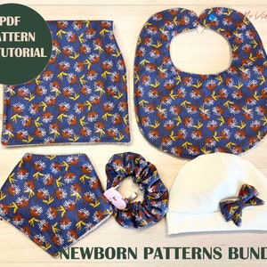 Peut inclure: Un ensemble de cinq patrons pour nouveau-nés dans un imprimé floral. L'ensemble comprend une bavette, un chapeau, un bandeau, une serviette à langer et une jupe. Les motifs sont tous dans un schéma de couleurs bleu et marron avec un motif floral blanc.