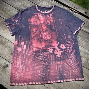 Puede incluir: Una camiseta azul marino con un diseño rosa y blanco. El diseño incluye una forma rectangular central con una espiral y otras formas, rodeada de motivos florales y de hojas rosas. Los bordes de las mangas y el cuello están adornados con un patrón rosa y blanco.