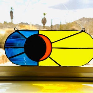 May include: A stained glass sun catcher with a blue, black, orange, and yellow design. The sun catcher is shaped like an oval and has a hanging loop at the top.