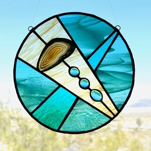 May include: A round stained glass window with a black border. The design features sections of teal, light blue, and cream-colored glass. A brown and tan agate stone is set in the center, with three small, round, teal glass accents.