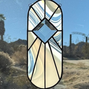 May include: A stained glass window hanging with a black border. The window has a central diamond shape and a design of cream, white, and blue glass. The window is hanging in front of a sunny outdoor scene.