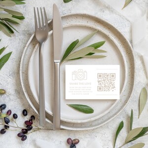 Può includere: Un biglietto bianco con un codice QR e il testo "SHARE THE LOVE" stampato su di esso. Il biglietto è posto su un piatto bianco con una forchetta e un coltello accanto.
