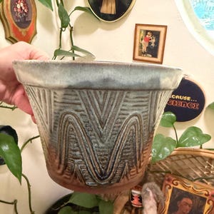 May include: A ceramic planter featuring a wide, flared rim and a tapered body. The exterior displays a textured, repeating geometric pattern of vertical zigzags in shades of blue, gray, and brown, with a smooth, light-colored glaze at the top.
