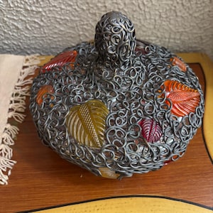May include: A decorative, round container with a metal filigree design. The container features colorful, leaf-shaped glass inserts in shades of red, orange, and yellow. The lid has a matching filigree handle.