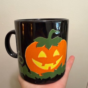 May include: Black ceramic mug featuring a smiling jack-o'-lantern design in orange, yellow, and green. The mug has a black handle and is held in a hand. A festive Halloween-themed item.