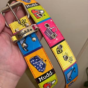 May include: A colorful Mudd belt with a silver buckle. The belt features a repeating pattern of various icons on a background of yellow, blue, pink, and green. The belt has the word "Mudd" printed on it.