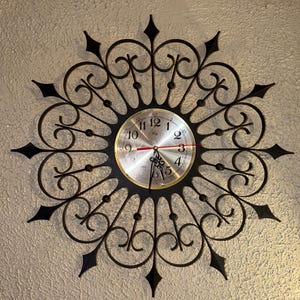 May include: A decorative wall clock with a black metal frame and a silver-colored clock face. The clock features a sunburst design with ornate scrollwork and pointed accents. The clock hands are black and red.