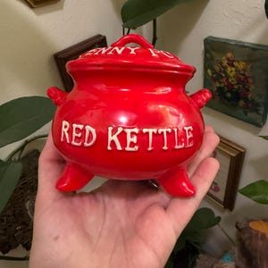 May include: A bright red ceramic cauldron-shaped pot with a matching lid. The text "RED KETTLE" is embossed in white on the front, and the text "PENNY POT" is embossed in white on the lid. The pot features three small legs and two side handles.
