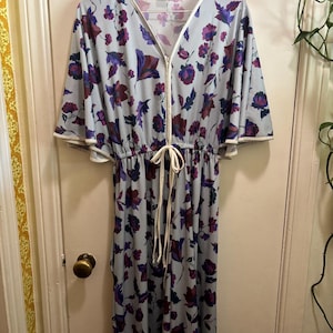 May include: A vintage-style, light blue dress with a floral pattern in shades of purple and burgundy. The dress features a V-neckline, a white zipper, and a drawstring waist. The sleeves are short and flared, with white trim.