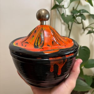 May include: A black ceramic bowl with a lid. The lid is painted with orange, yellow, and black swirls and has a silver knob. The bowl has a glossy finish with orange drips down the side. The bowl is held by a hand.