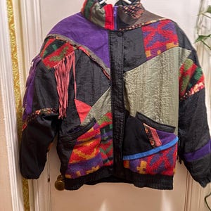 May include: A colorful patchwork jacket with a high collar, featuring a mix of textures and patterns. The jacket includes black, purple, red, and green panels, with fringe accents and a zippered front. The jacket is hanging on a white hanger.
