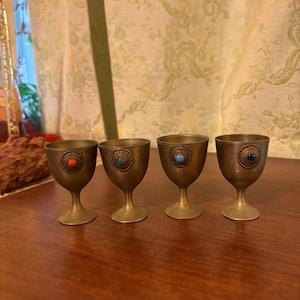 May include: Four small brass goblets with a decorative gemstone in the center of each. The gemstones are red, green, turquoise, and blue. The goblets have a rounded bowl shape and a slender stem.
