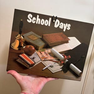 May include: A vintage vinyl record album cover with a brown background and the text "School Days" in white. The cover features a still life of school supplies, including a gold bell, a red apple, books, a graduation cap, and a diploma.