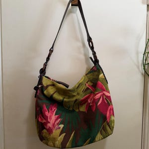 May include: A shoulder bag with a tropical floral print. The bag features a dark brown leather strap with wooden accents. The main body of the bag is a mix of green, brown, and pink floral patterns. The bag is hanging on a gold hook.