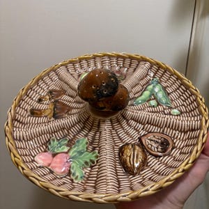May include: A brown ceramic serving dish shaped like a woven basket with a mushroom-shaped handle. The dish has six sections, each decorated with a different fruit or vegetable, including mushrooms, peas, walnuts, and turnips.