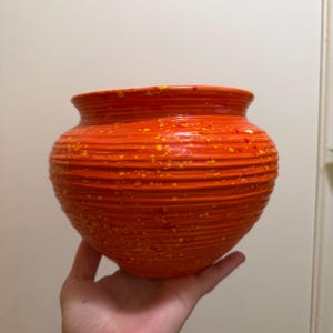 May include: A round, orange ceramic planter featuring a horizontal ribbed texture across its surface. The exterior is decorated with a speckled pattern of small yellow and red paint flecks, creating a vibrant, multi-colored appearance.